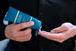  A man in a black shirt holding a Manuka Biotic Light Day Cream bottle, squeezing a small amount onto his finger to apply to his rosacea-prone skin.