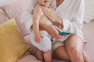  An adorable baby wearing a diaper stands on their feet, supported by their mother's loving arms. In her other hand, the mother holds a tube of Manuka Biotic Eczema Relief Body Lotion, which she has applied to the baby's skin on his chest