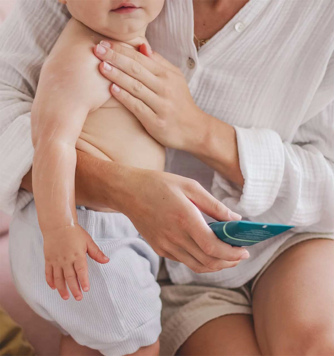  A woman holding a teal green bottle is sitting on a blush pink couch while she holds a baby upright with one of her hands on the baby's chest, suggesting that she is applying lotion.  