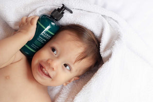  An adorable, happy baby with a bright smile, holding a bottle of Eczema Relief Body Lotion near their face.